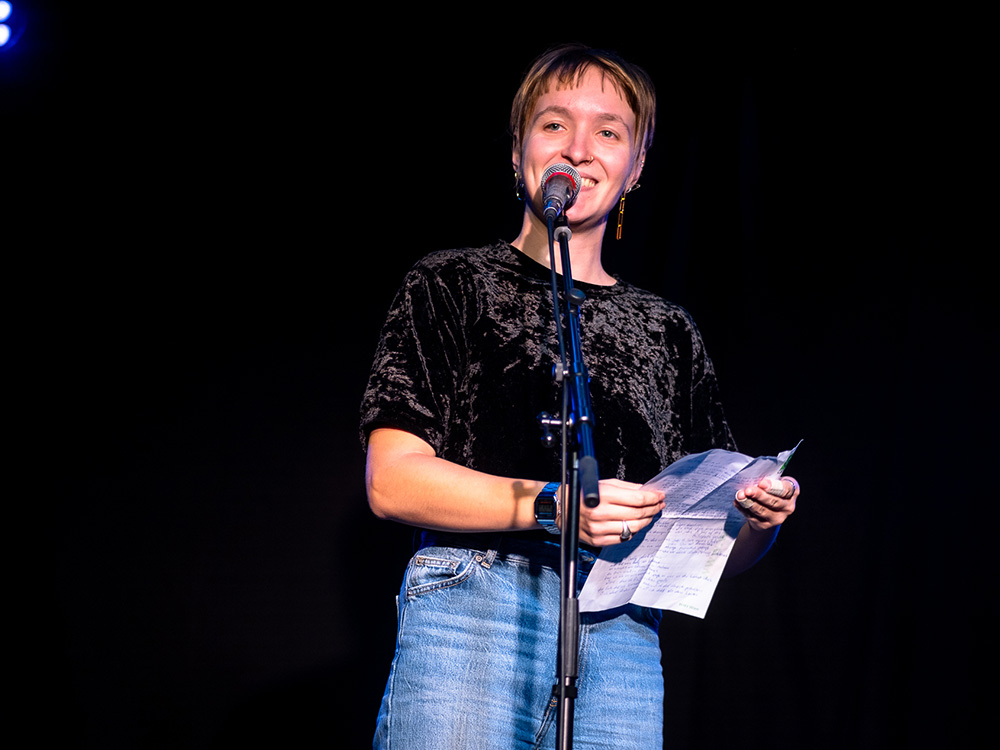 Eine junge Frau mit kurzen blonden Haaren steht auf einer Bühne, hält einen Zettel in der Hand, spricht in ein Mikrofon und lächelt in die Kamera.
