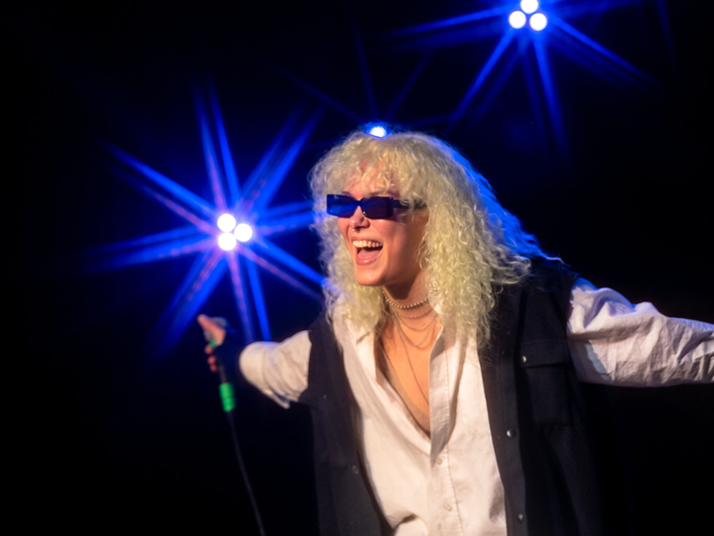 Junge blonde Frau mit Sonnenbrille lacht und singt auf eine Bühne.