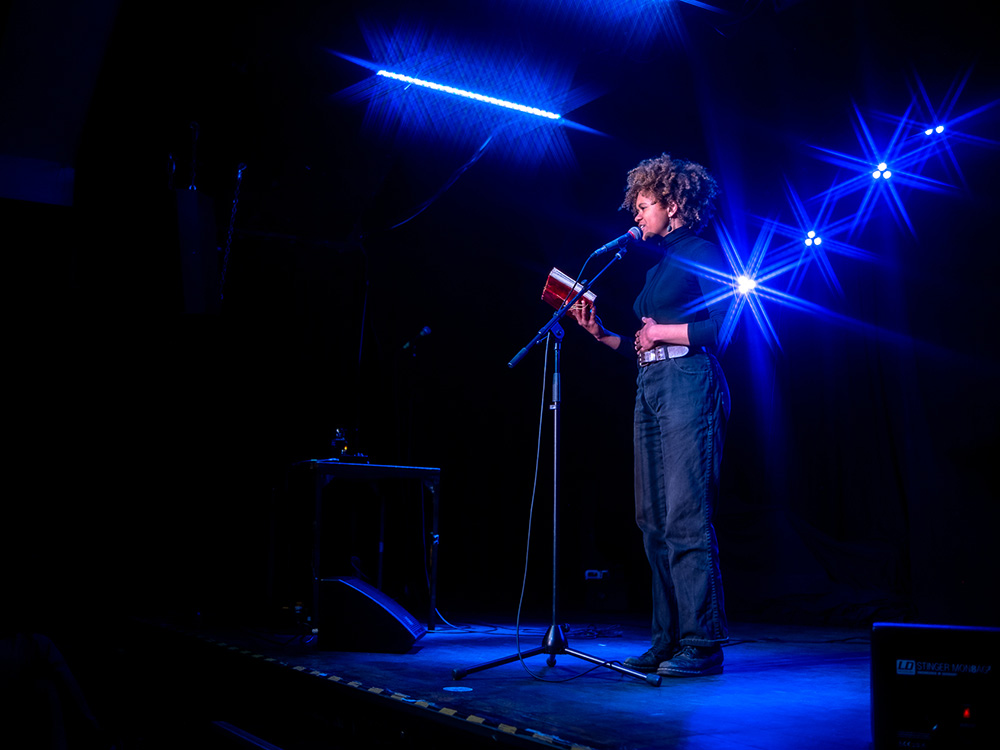 Junge Frau steht auf einer Bühne mit einem roten Notizheft in der Hand und spricht in ein Mikrofon.