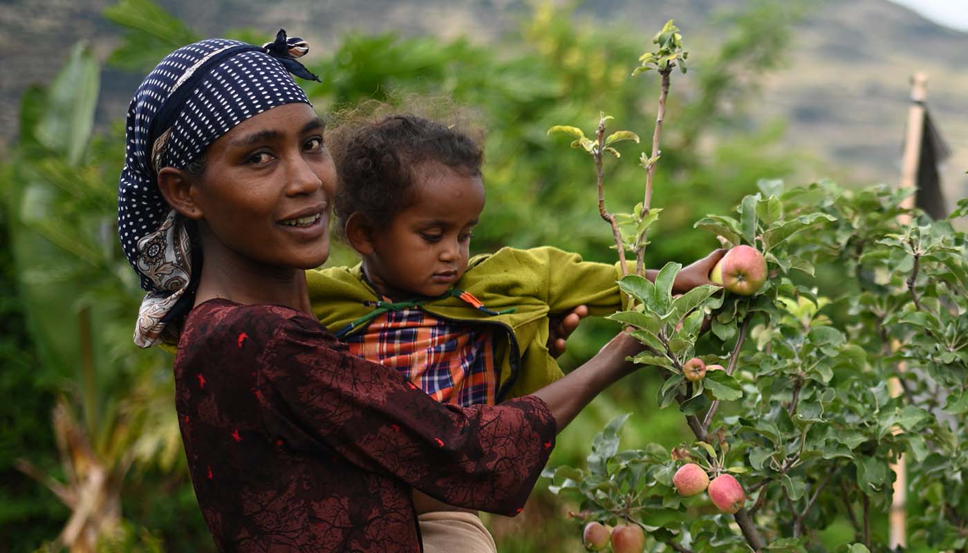 Eine Mutter mit Kind steht neben einem kleinen Apfelbaum