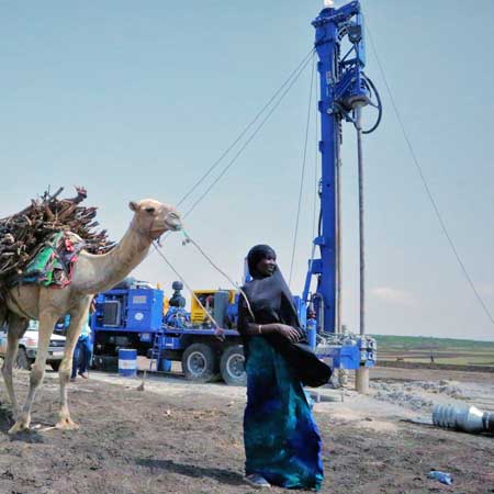 Frau geht mit Kamel an einem Wasserbohrgerät vorbei