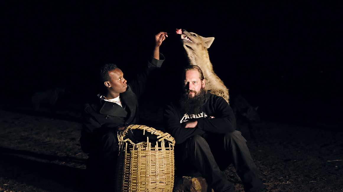 Mann sitzt vor Hyäne die gerade von einem Äthiopier gefüttert wird