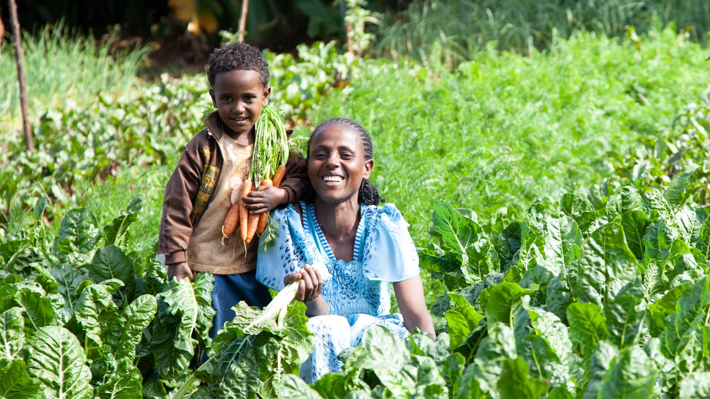 Eine äthiopische Frau und ihr Sohn sitzen in einem Gartenbeet und halten lächelnd Karotten hoch