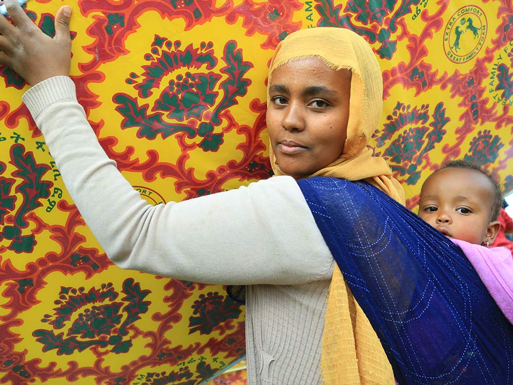 Äthiopische Frau mit Baby am Rücken trägt eine bunte Matratze