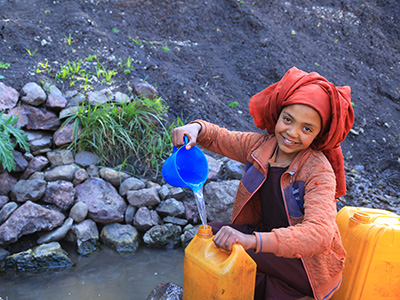 Äthiopisches Mädchen mit rotem Tuch am Kopf schöpft Wasser aus einem Tümpel in einen gelben Wasserkanister.