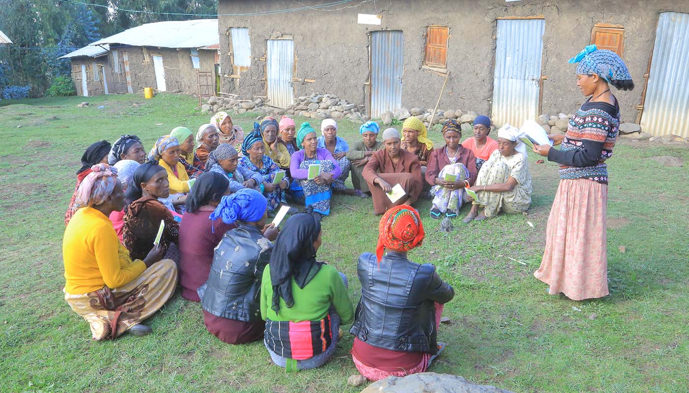 Eine Gruppe Frauen sitzt beim Mikrokredit-Training in einem Halbkreis am Boden. Die Vortragende steht und liest aus einem Buch vor