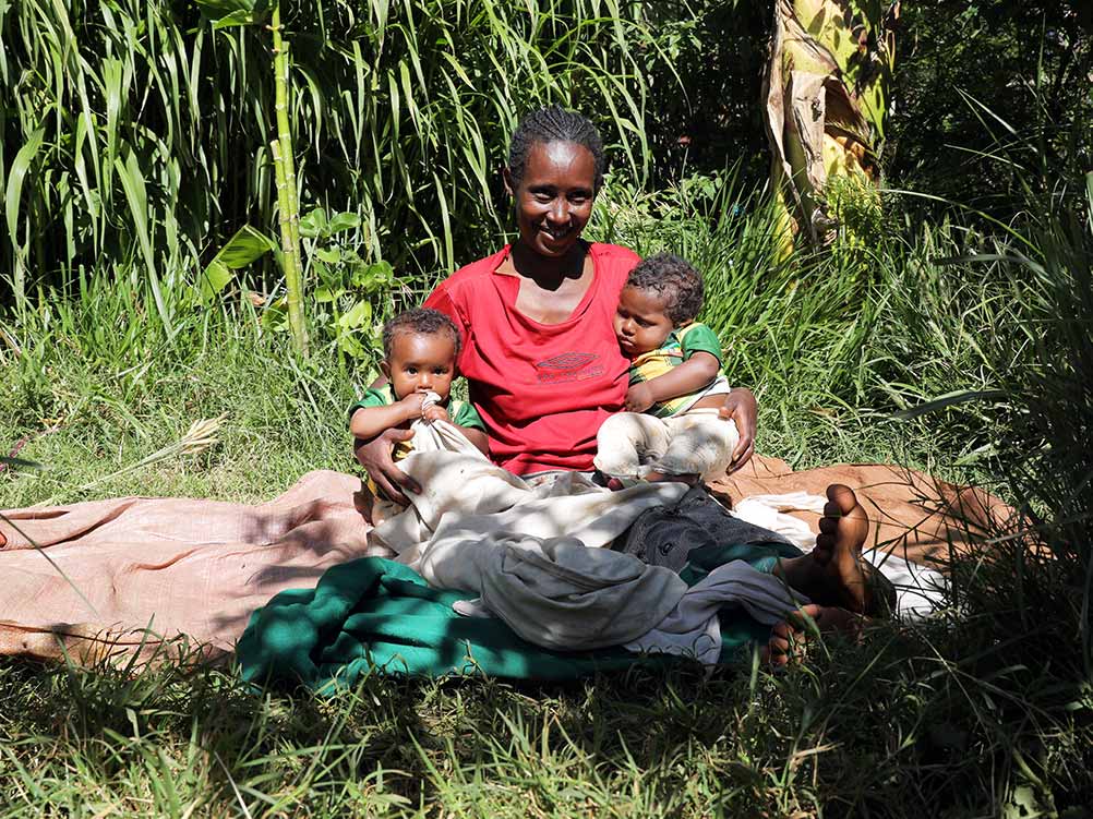 Ein Mutter sitzt mit ihren Zwillingen auf einer Decke in einer Wiese.