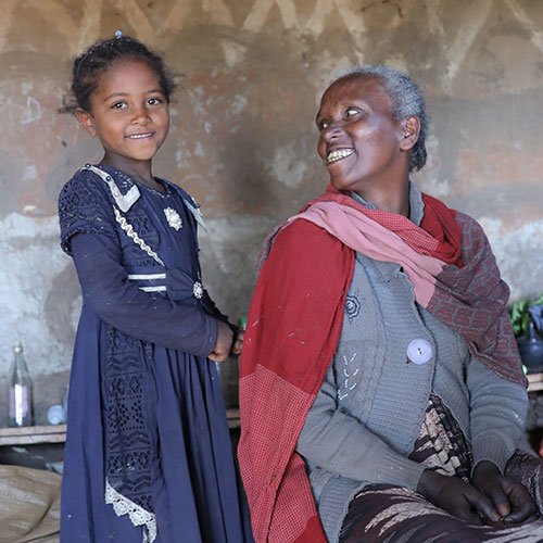 Junges äthiopisches Mädchen steht und lächelt in die Kamera. Eine ältere Frau sitzt neben ihr und sieht sie freundlich an.