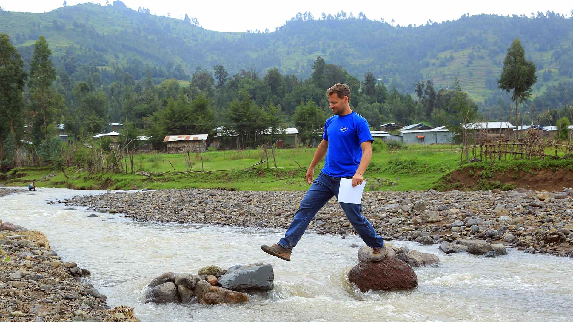 Markus Schwarz-Herda überquert Flusslauf in Äthiopien, indem er von Stein zu Stein springt. Er trägt ein blaues T-Shirt und hält Projektunterlagen in der Hand; im Hintergrund ist eine hügelige Landschaft zu sehen.