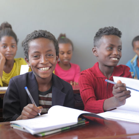 Ein Junge lächelt in die Kamera, während er in einem Klassenzimmer in sein Notizbuch schreibt.