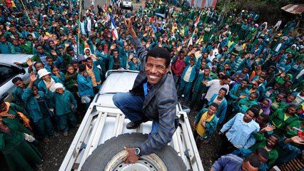 Haile Gebrselassie steht auf einem weißen Geländewagen und hebt triumphierend die Faust, während eine große Menschenmenge ihn jubelnd umringt.