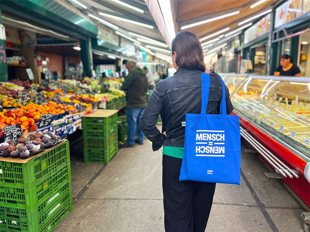 Eine Frau geht über den Wochenmarkt, ihre Stofftasche trägt den Aufdruck „Mensch ist gleich Mensch“.