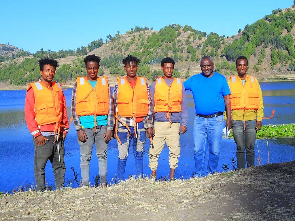 Fünf Männer in orangefarbenen Schwimmwesten und ein Mann in blauem Hemd stehen auf einer Böschung am Mahyibar-See in Albuko.