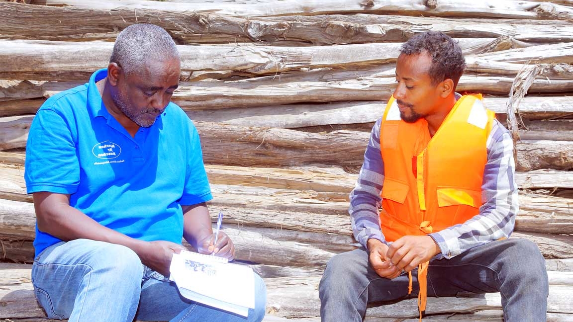 Zwei Männer sitzen auf Holzstämmen. Einer schreibt in einen Notizblock, der andere hört ihm aufmerksam zu.