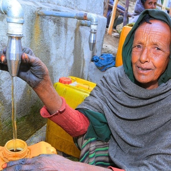 Eine Frau sitzt an einer Quellfassung und füllt sauberes Wasser in ihren gelben Kanister.