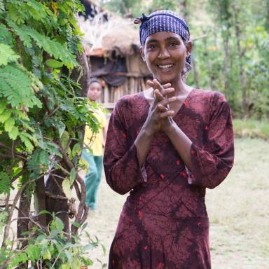 Nahaufnahme einer lächelnden jungen Frau in einem gemusterten Kleid und mit einem Kopftuch, die die Hände vor der Brust hält. Im Hintergrund sind undeutlich ein Kind und eine Hütte zu sehen, umgeben von grüner Vegetation.