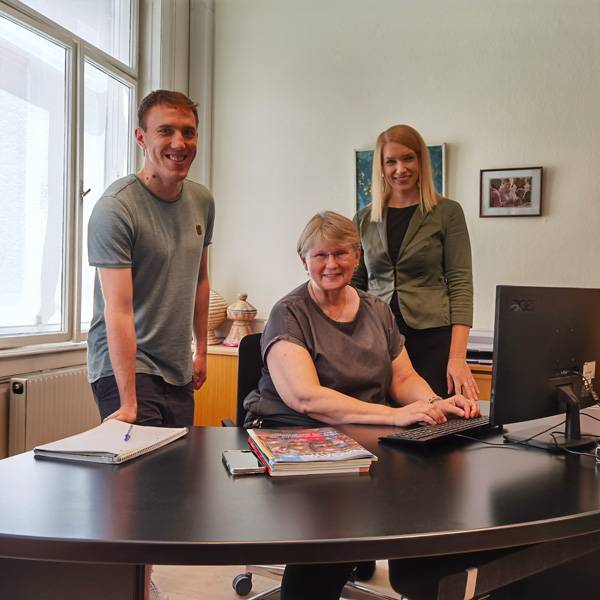Ein Bild vom Spendenservice-Team von Menschen für Menschen. Zwei Frauen und ein Mann sitzen bzw. stehen an einem Schreibtisch und lächeln in die Kamera