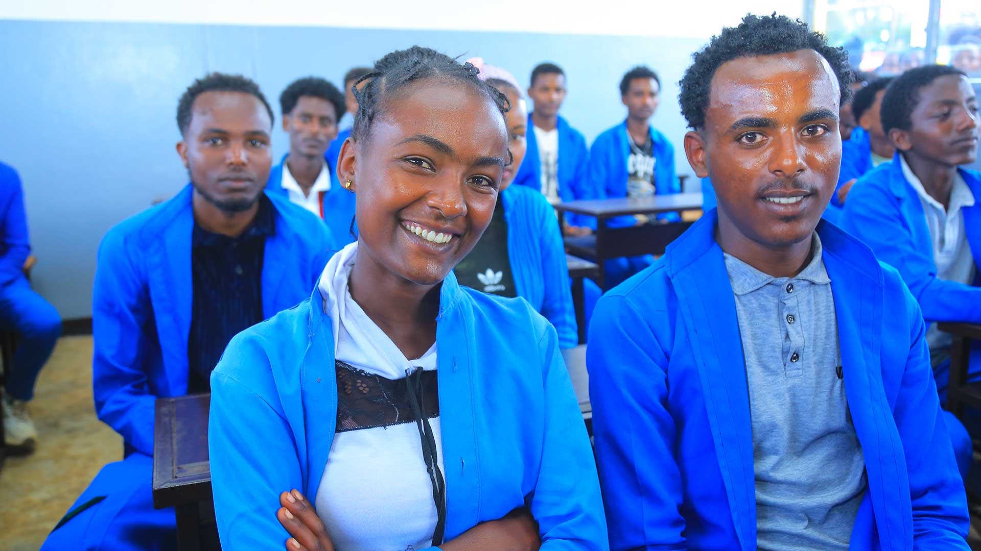 Kinder in blauen Schuluniformen sitzen auf Bänken im Klassenzimmer und lächeln in die Kamera.