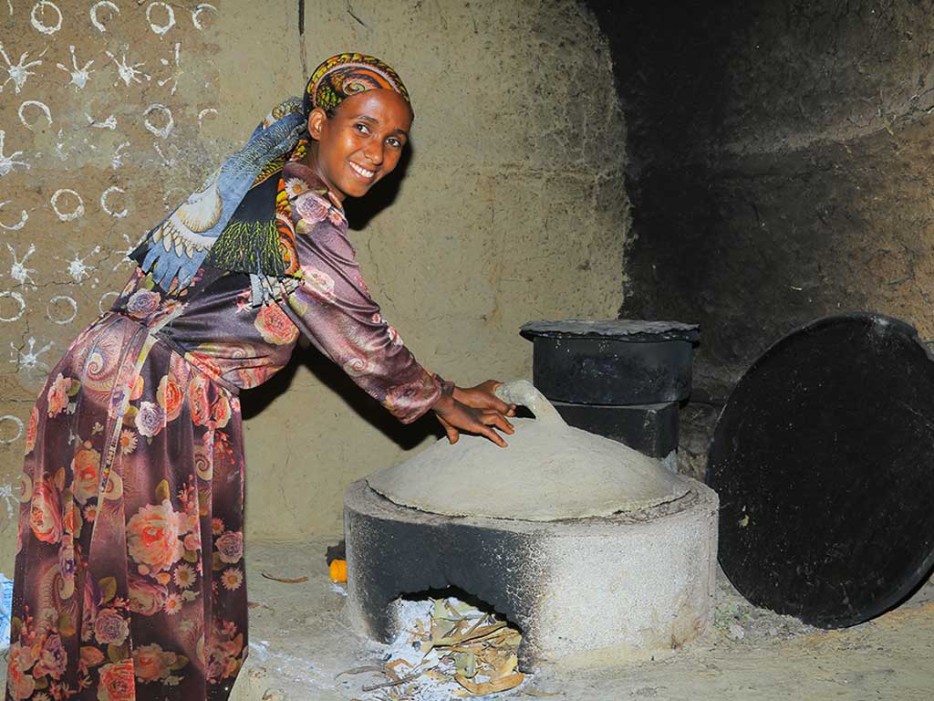 Eine Frau in traditioneller Kleidung bereitet in einer einfachen Küche Injera auf einem holzsparenden Zementofen zu.