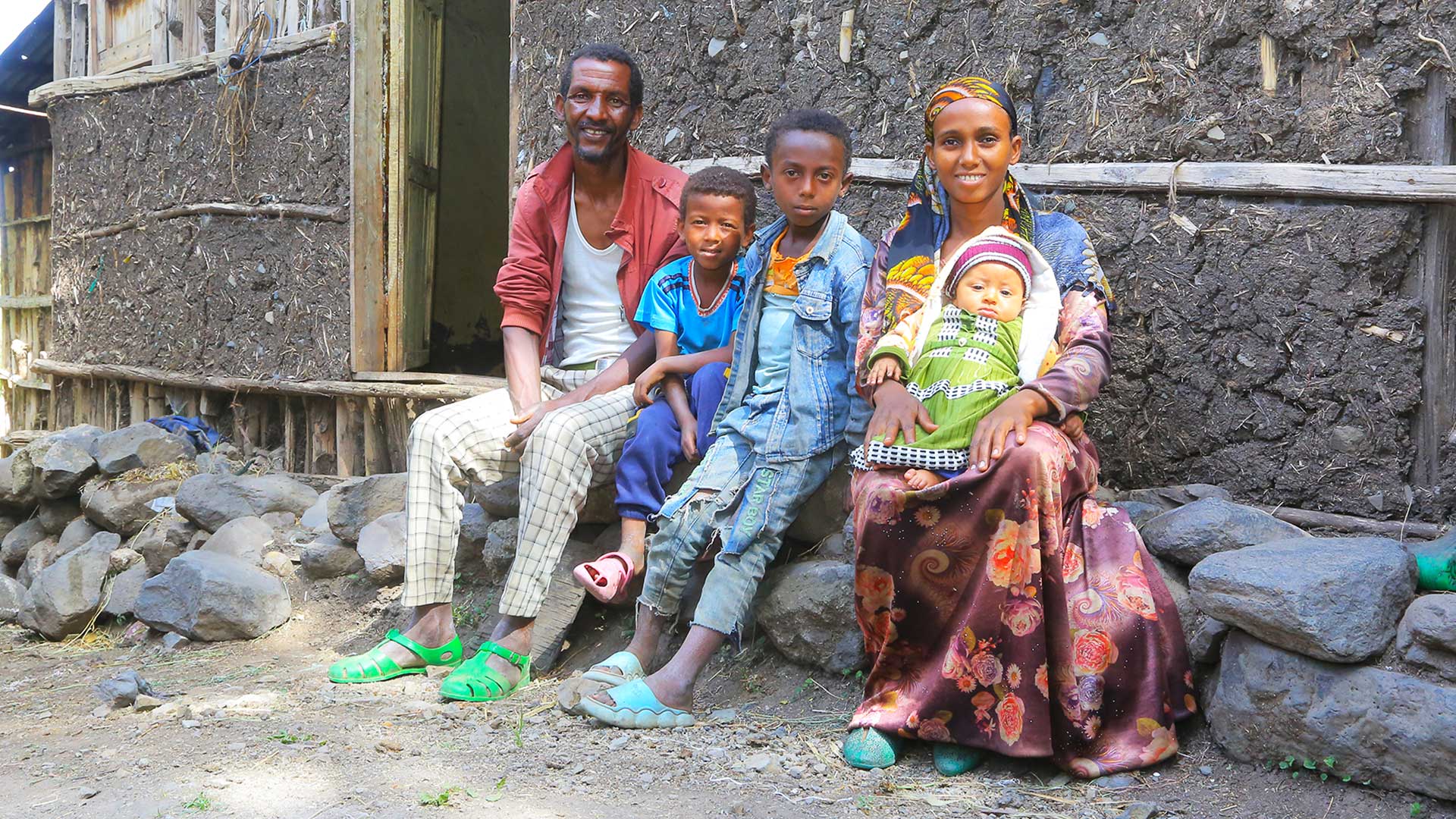 Eine Familie mit zwei Erwachsenen und drei Kindern sitzt vor einem einfachen Haus mit einer mit Lehm verputzten Wand.