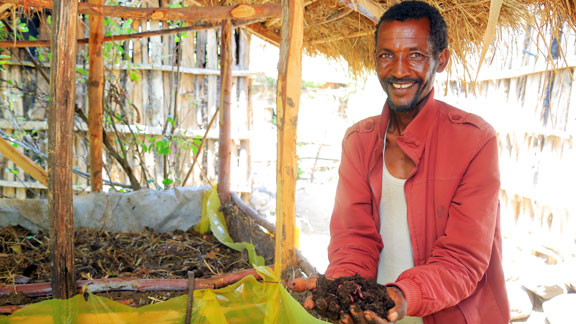 Ein Mann mit rotem Hemd hält lächelnd fruchtbare Erde aus einem Wurmbeet in den Händen.