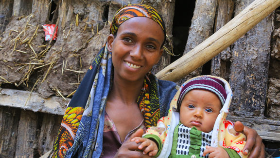 Eine lächelnde Frau sitzt vor ihrem Lehmhaus und hält ihr Baby mit Mütze liebevoll im Arm.