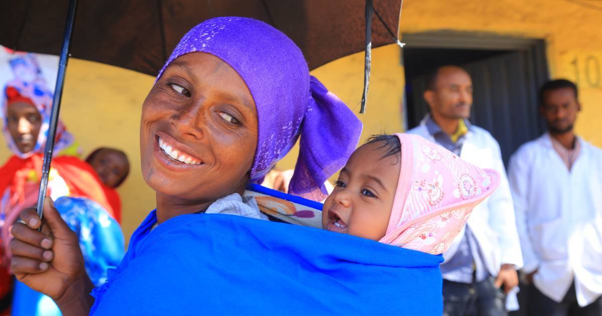Eine lächelnde Frau mit lila Kopftuch und leuchtend blauem Tragetuch trägt ein Baby mit einer rosa Mütze auf dem Rücken, während sie einen schwarzen Regenschirm hält. Im verschwommenen Hintergrund stehen mehrere Männer vor einem gelben Gebäude.