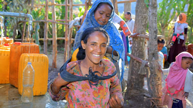 Eine Frau hilft der anderen, den frisch mit Wasser gefüllten Kanister auf ihren Rücken zu befestigen.