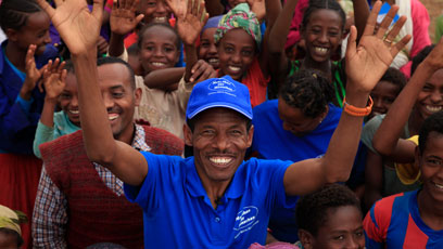 Haile Gebrselassie steht im Vordergrund, die Arme jubelnd erhoben, umgeben von einer Gruppe lächelnder Kinder.