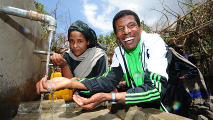 Haile Gebrselassie steht gemeinsam mit einer Frau an einer Quellfassung und wäscht sich die Hände.