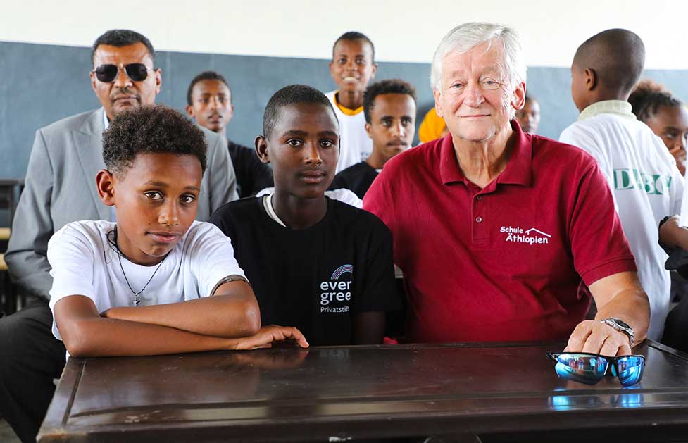 Peter Krasser sitzt mit Kindern auf einer Schulbank im Klassenzimmer.