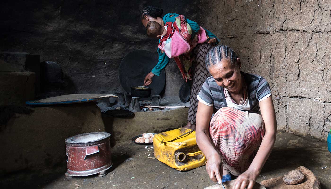 Zwei äthiopische Frauen. Eine im Vordergrund, die Zwiebeln schneidet und eine im Hintergrund mit einem Baby im Arm, die vor einer traditionellen Feuerstelle steht.