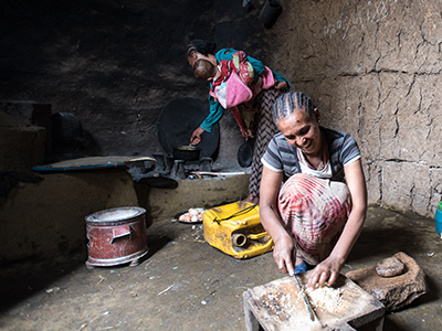 zwei äthiopische Frauen bei der Hausarbeit. Eine im Vordergrund schneidet Zwiebeln auf einem Brett und im Hintergrund hält eine Frau ein Baby im Arm und blickt auf eine traditionelle Feuerstelle