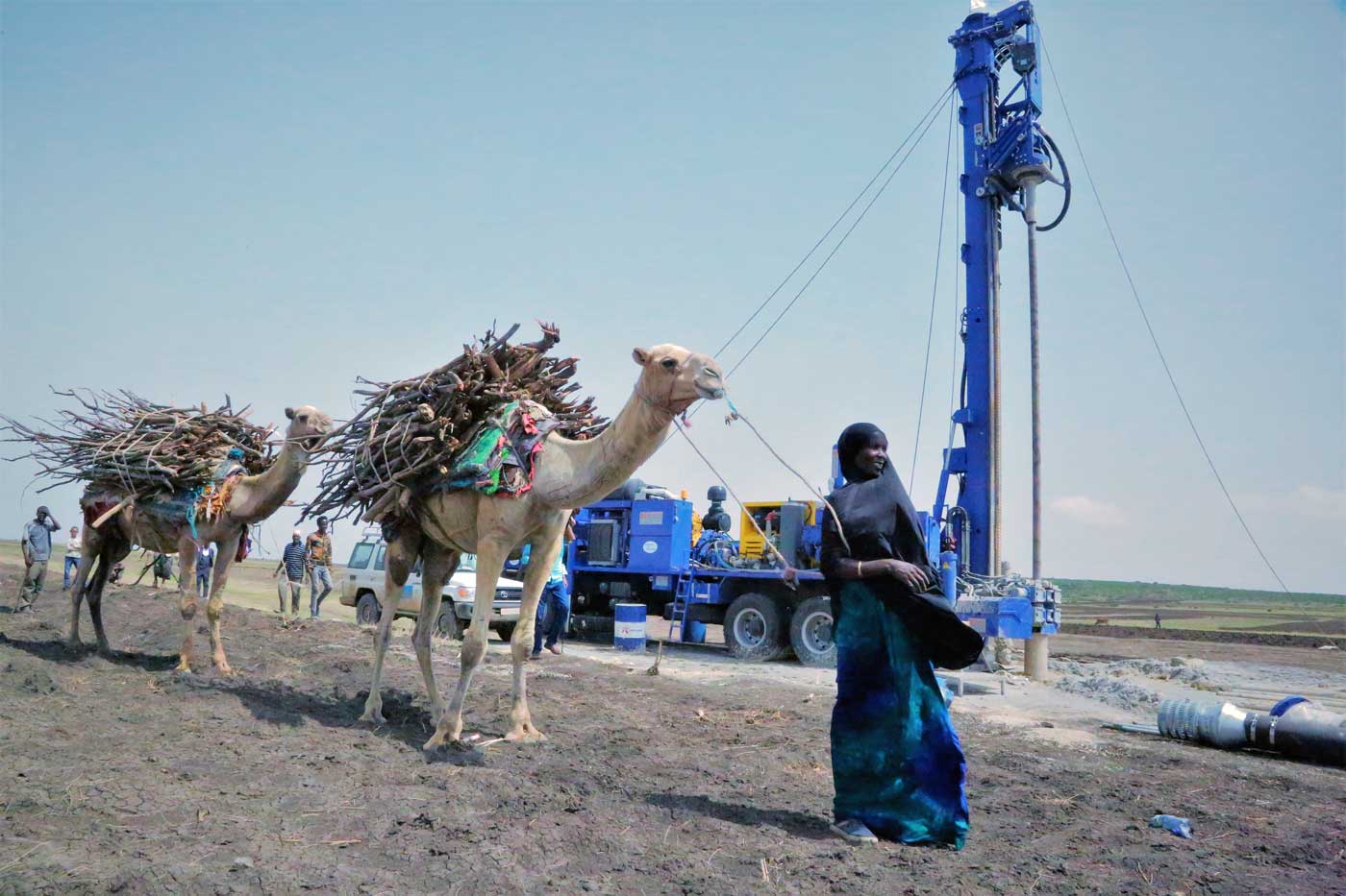 Eine Frau geht mit Kamelen mit Holz am Rücken an einem Brunnenbohrgerät vorbei