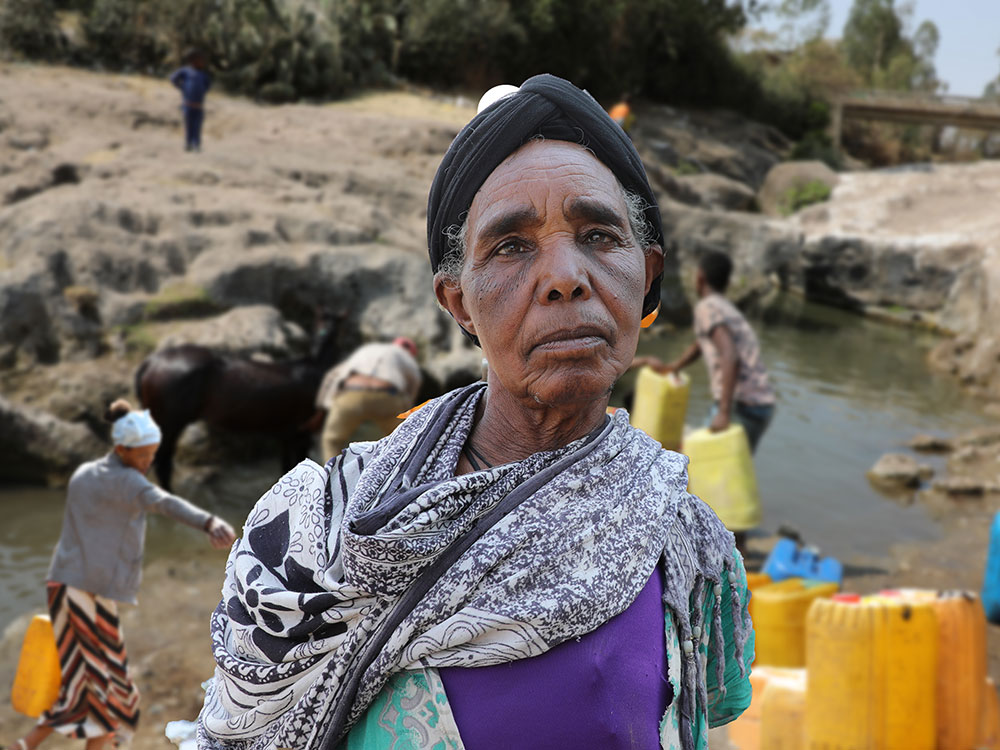 Eine äthiopische Frau steht vor einem Fluss aus dem im Hintergrund Wasser geschöpft wird