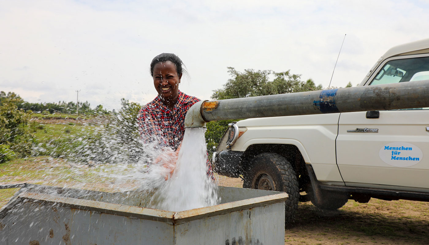 Eine lachende Frau steht an einem Wasserrohr aus dem viel Wasser sprudelt