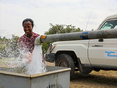 Eine lachende Frau steht an einem Wasserrohr aus dem viel Wasser sprudelt