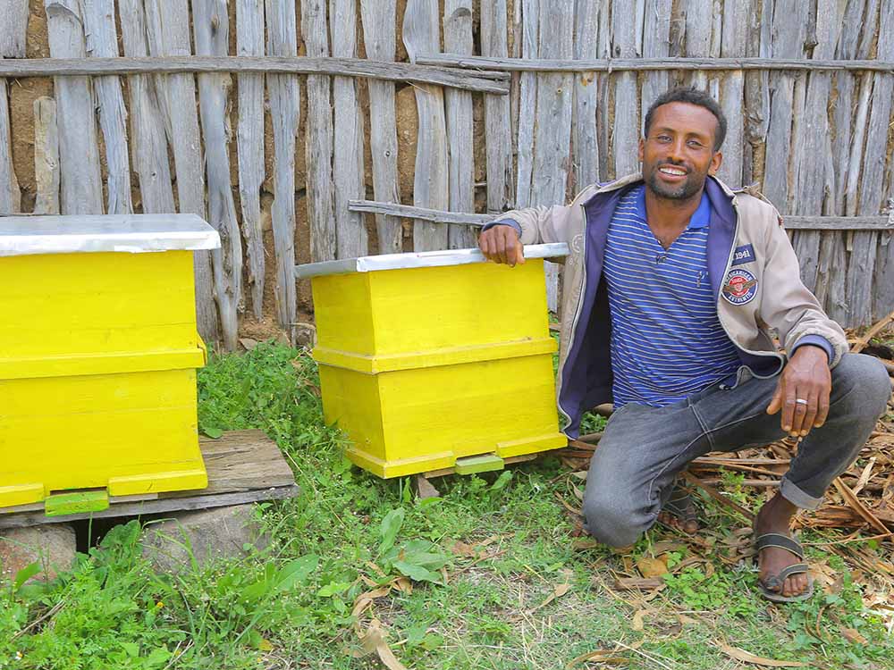Äthiopischer Mann lächelt in die Kamera. Er hockt neben zwei modernen gelben Bienenstöcken und lehnt sich an einem an.