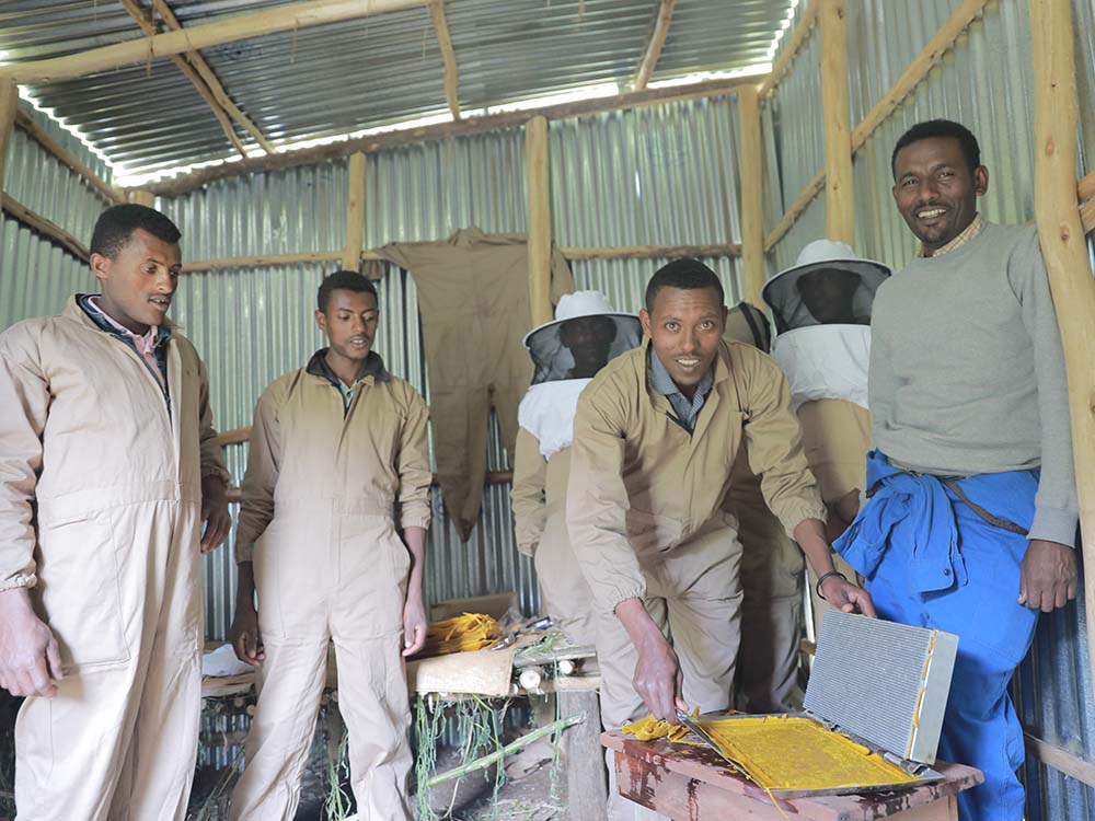 1 junger äthiopischer Mann stellt Rähmchen aus Wachs für Bienen her und lächelt dabei in die Kamera. 5 weitere Männer stehen hinter ihm und neben ihm und sehen ihm dabei zu.