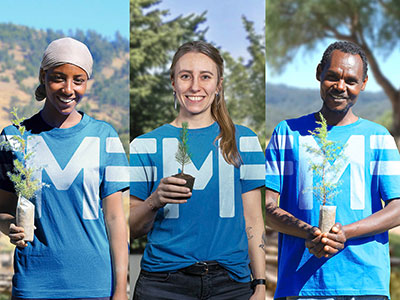 Drei Menschen aus unterschiedlichen Herkunftsländern stehen nebeneinander im Freien und halten junge Bäumchen in den Händen. Sie tragen blaue T-Shirts mit einem großen M 