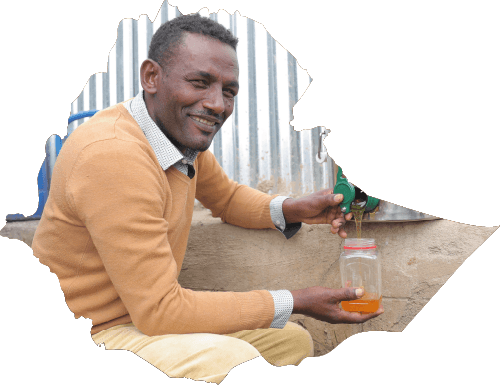 Äthiopischer Mann blickt in die Kamera und lächelt. Er hält ein Glas in der Hand und befüllt es über einen silbernen Behälter mit Honig.